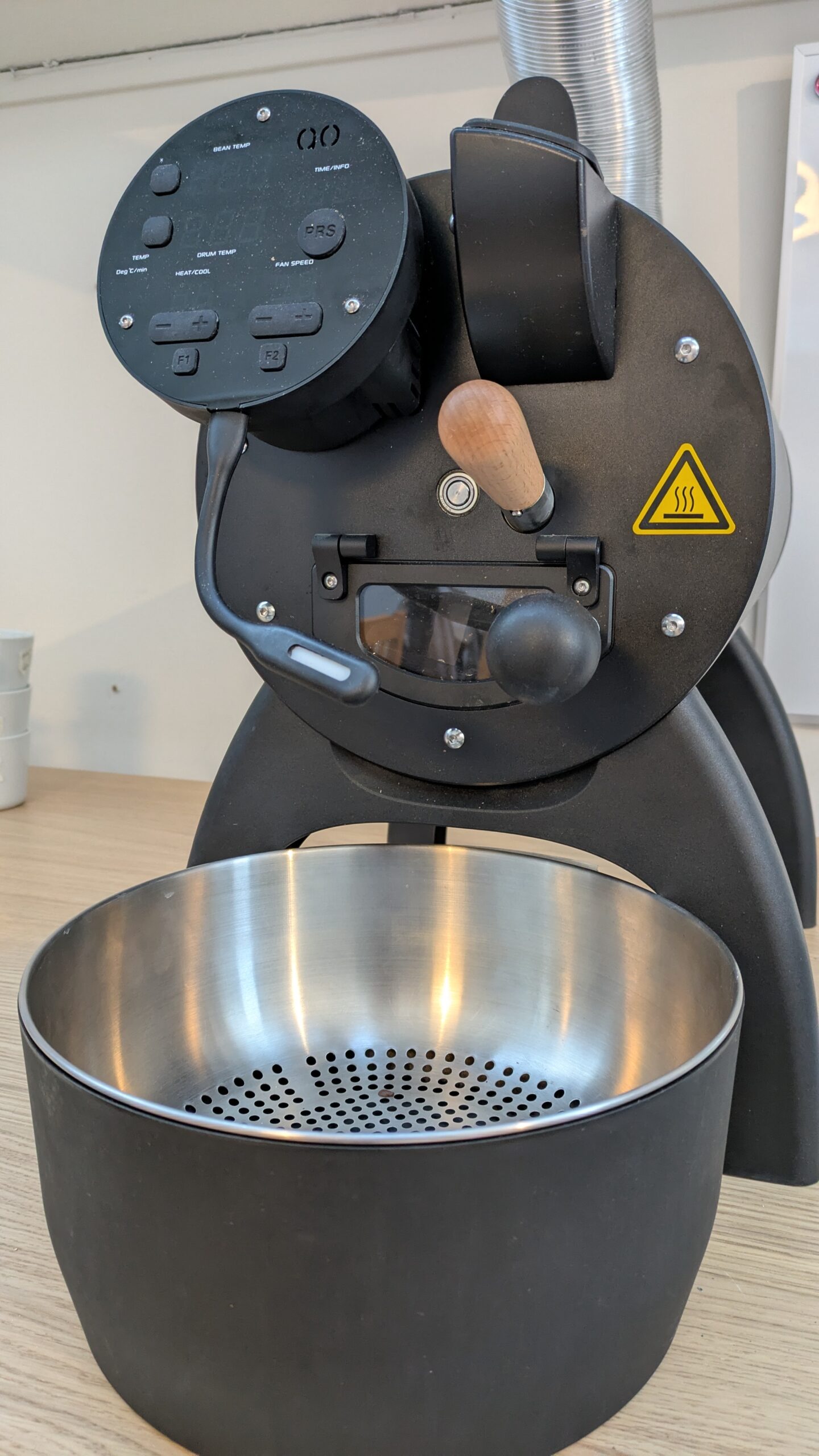 Close-up of a coffee roaster machine (Aillio Bullet R1) featuring controls for temperature and timer settings, with a cooling traz at the bottom.