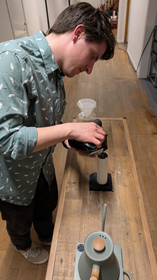 Pouring beans into a Coffee grinder. A Brewing space full of coffee equipment like the Fellow Stagg Pour over kettle and hario v60. The Scale Is a Timemore Blackmirror Nano and the Grinder a Timemore Chestnut c3.